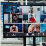 A large monitor in a modern office displays a nine-person video conference grid, with each participant in a different indoor setting, while a person in the foreground watches the screen.