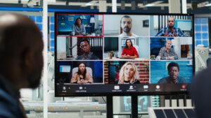 A large monitor in a modern office displays a nine-person video conference grid, with each participant in a different indoor setting, while a person in the foreground watches the screen.