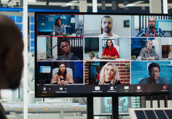 A large monitor in a modern office displays a nine-person video conference grid, with each participant in a different indoor setting, while a person in the foreground watches the screen.