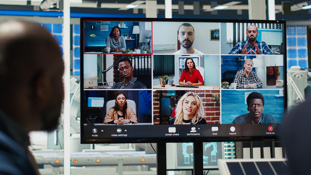 A large monitor in a modern office displays a nine-person video conference grid, with each participant in a different indoor setting, while a person in the foreground watches the screen.