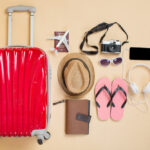 Flat lay of travel essentials including a red suitcase, straw hat, flip‑flops, camera, headphones, smartphone, passport, and notebook arranged on a beige background.