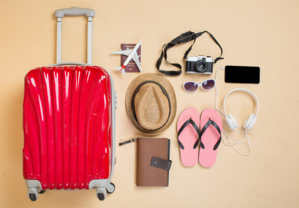 Flat lay of travel essentials including a red suitcase, straw hat, flip‑flops, camera, headphones, smartphone, passport, and notebook arranged on a beige background.