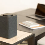 Rectangular black wireless computer speaker with fabric sides on a small wooden desk shelf in a modern home office, with an open laptop and notebooks blurred in the background.