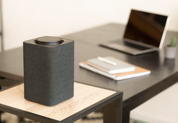 Rectangular black wireless computer speaker with fabric sides on a small wooden desk shelf in a modern home office, with an open laptop and notebooks blurred in the background.
