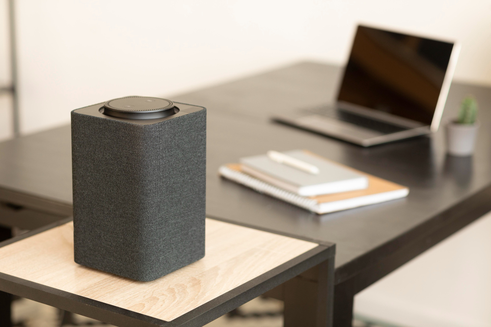Rectangular black wireless computer speaker with fabric sides on a small wooden desk shelf in a modern home office, with an open laptop and notebooks blurred in the background.