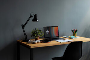A simple wooden desk against a gray wall holds a laptop, desk lamp, plant, notebooks, and a cup of colorful pens, with a black chair in front.