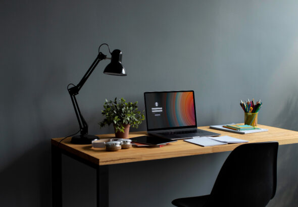 A simple wooden desk against a gray wall holds a laptop, desk lamp, plant, notebooks, and a cup of colorful pens, with a black chair in front.