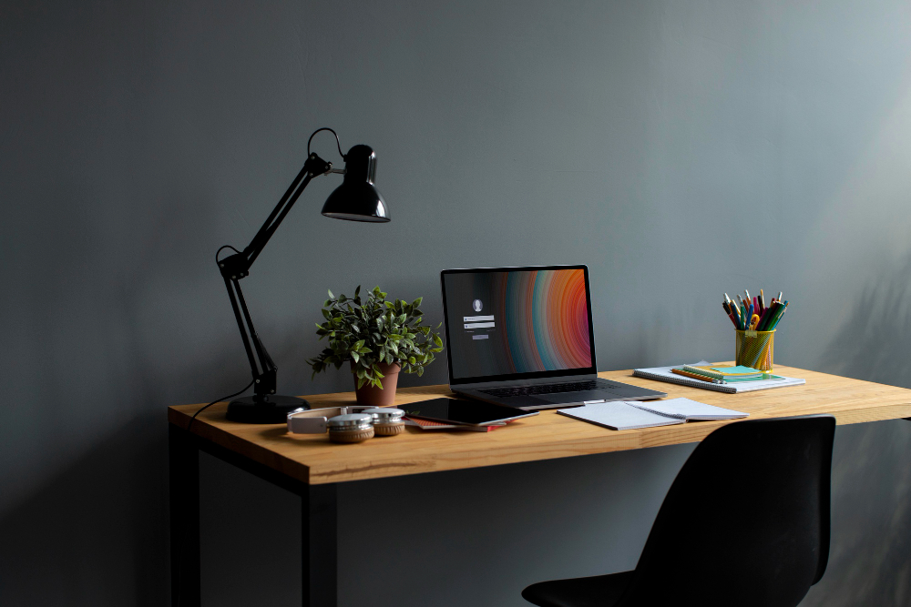 A simple wooden desk against a gray wall holds a laptop, desk lamp, plant, notebooks, and a cup of colorful pens, with a black chair in front.