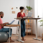 Two people working in a bright home office, one using a laptop at a standing desk and the other seated on an exercise ball at a desk.