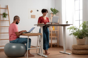 Two people working in a bright home office, one using a laptop at a standing desk and the other seated on an exercise ball at a desk.