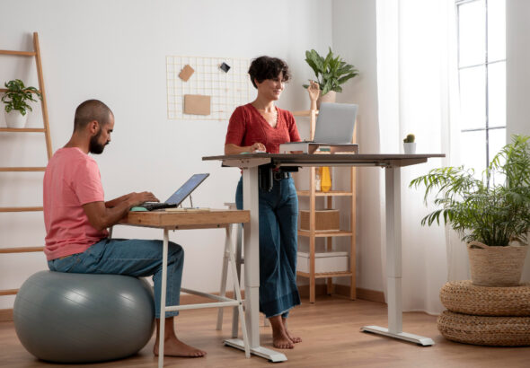 Two people working in a bright home office, one using a laptop at a standing desk and the other seated on an exercise ball at a desk.