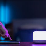 Hand tapping a smartphone on a desk beside a glowing smart light in a dim, modern room.