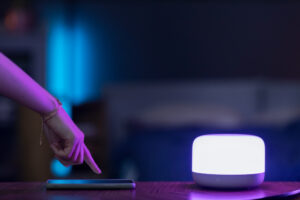 Hand tapping a smartphone on a desk beside a glowing smart light in a dim, modern room.