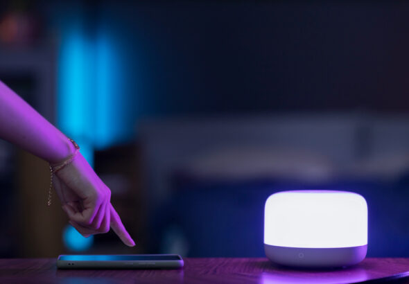 Hand tapping a smartphone on a desk beside a glowing smart light in a dim, modern room.
