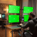 Man working at a desk with multiple monitors mounted on adjustable arms, displaying green screens.