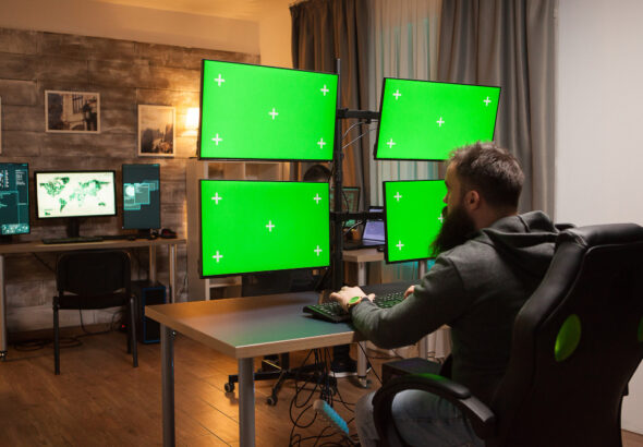 Man working at a desk with multiple monitors mounted on adjustable arms, displaying green screens.