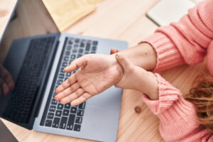 Person holding their wrist in discomfort while using a laptop on a desk.