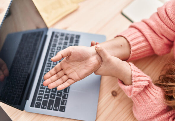 Person holding their wrist in discomfort while using a laptop on a desk.