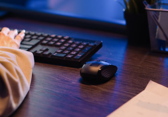 A person working at a computer with one hand on a keyboard and an ergonomic wireless mouse placed on a dark wooden desk in a softly lit office.