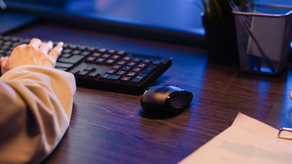 A person working at a computer with one hand on a keyboard and an ergonomic wireless mouse placed on a dark wooden desk in a softly lit office.