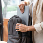 Person placing a laptop into a gray backpack near a window
