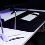 White dual-antenna Wi-Fi router on a minimalist desk setup with keyboard, mouse, and monitor in a dimly lit home office.