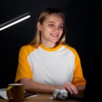 Young woman coding on a laptop at night under a modern desk lamp.