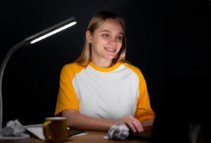 Young woman coding on a laptop at night under a modern desk lamp.