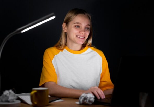 Young woman coding on a laptop at night under a modern desk lamp.