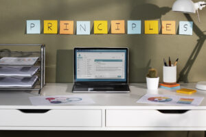 Minimalist desk workspace with laptop, desk organizer trays, lamp, cactus, and colorful sticky notes on the wall spelling “PRINCIPLES.”