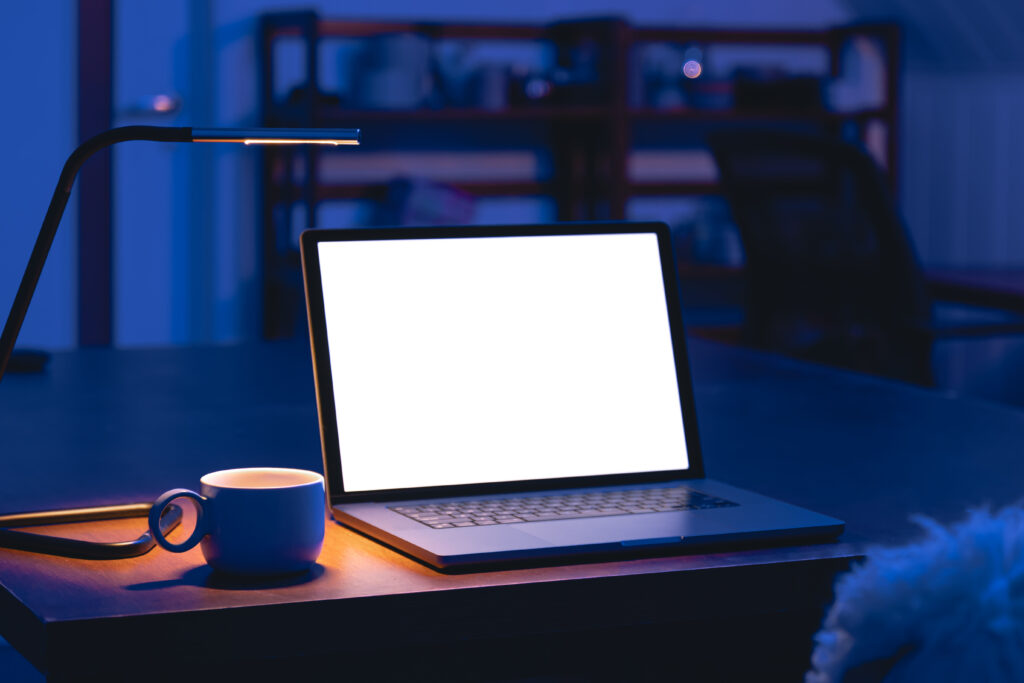 An open laptop with a bright blank white screen sits on a wooden table beside a coffee mug and a slim desk lamp in a dark blue-lit room.