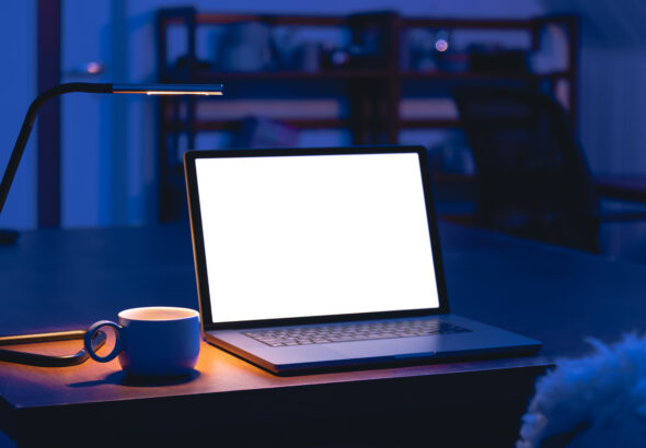 An open laptop with a bright blank white screen sits on a wooden table beside a coffee mug and a slim desk lamp in a dark blue-lit room.