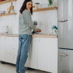 Woman washing dishes in a bright modern kitchen while a robot vacuum cleans the floor nearby.