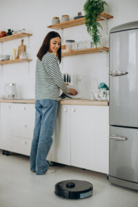 Woman washing dishes in a bright modern kitchen while a robot vacuum cleans the floor nearby.
