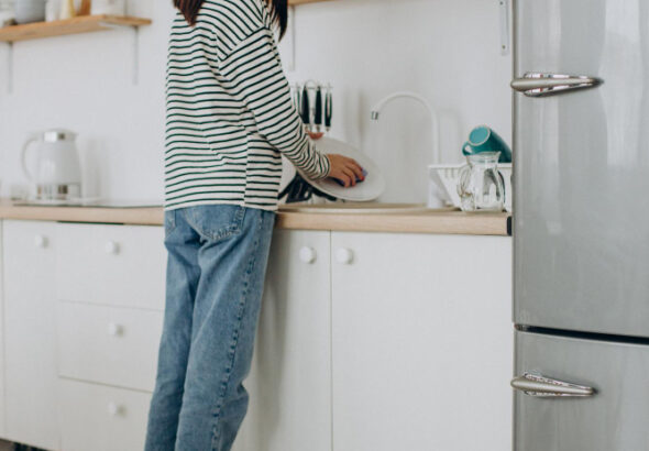 Woman washing dishes in a bright modern kitchen while a robot vacuum cleans the floor nearby.