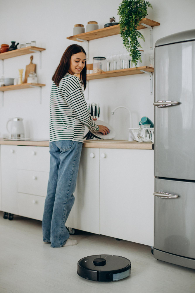 Woman washing dishes in a bright modern kitchen while a robot vacuum cleans the floor nearby.