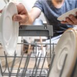 Person loading white plates into the lower rack of a dishwasher in a home kitchen.